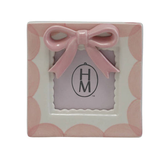 A square ceramic picture frame with a scalloped pink border, a pink bow on top, and a central scalloped window displaying a monogram logo.