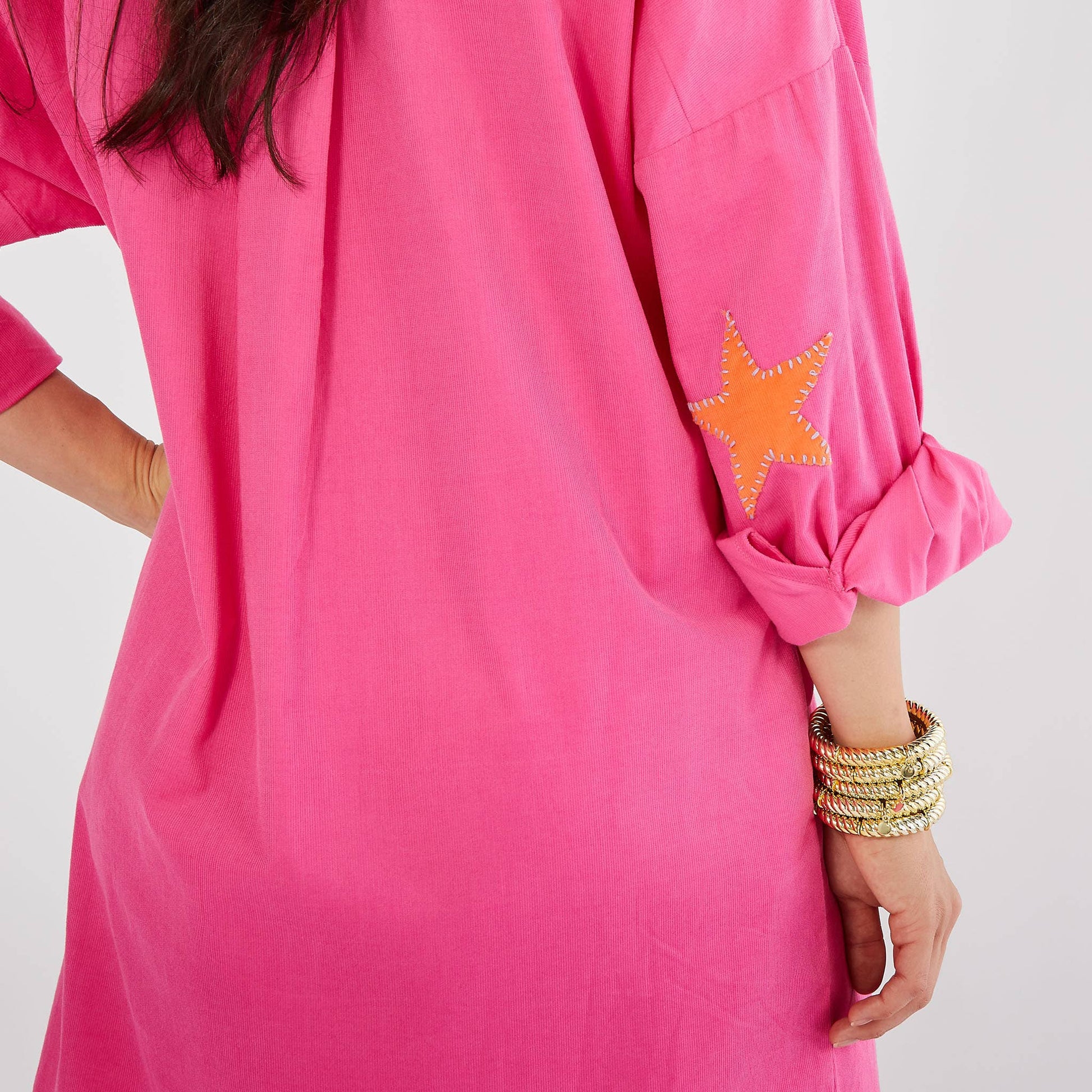 A close-up of a bright pink tunic dress with a rolled-up sleeve featuring an orange star patch with white stitching, worn by a model with stacked gold bracelets.