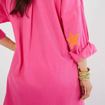 A close-up of a bright pink tunic dress with a rolled-up sleeve featuring an orange star patch with white stitching, worn by a model with stacked gold bracelets.
