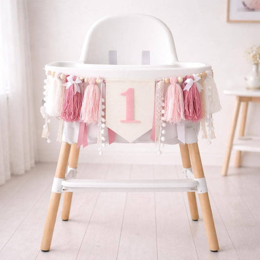 A white high chair with light wood legs decorated with pink tassels, lace trim, and a central banner featuring the number 1, set in a bright nursery.