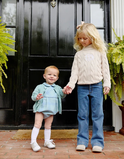 Two young girls standing on a brick porch in front of a black front door, holding hands. The older girl wears a cream cable-knit turtleneck sweater with a white floral applique, blue jeans, and cream shoes.