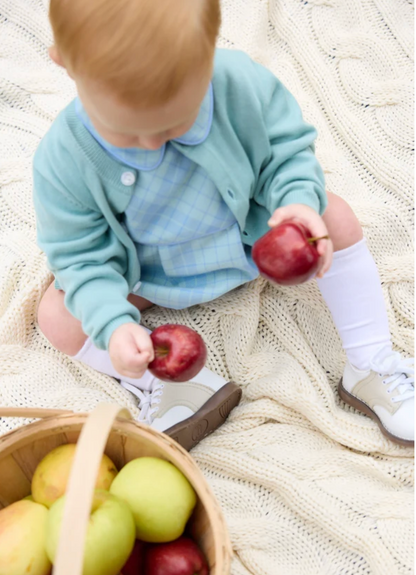 A baby sitting on a cream cable-knit blanket, wearing a light blue plaid shirt and a mint green cardigan, holding red apples next to a wooden basket filled with apples.