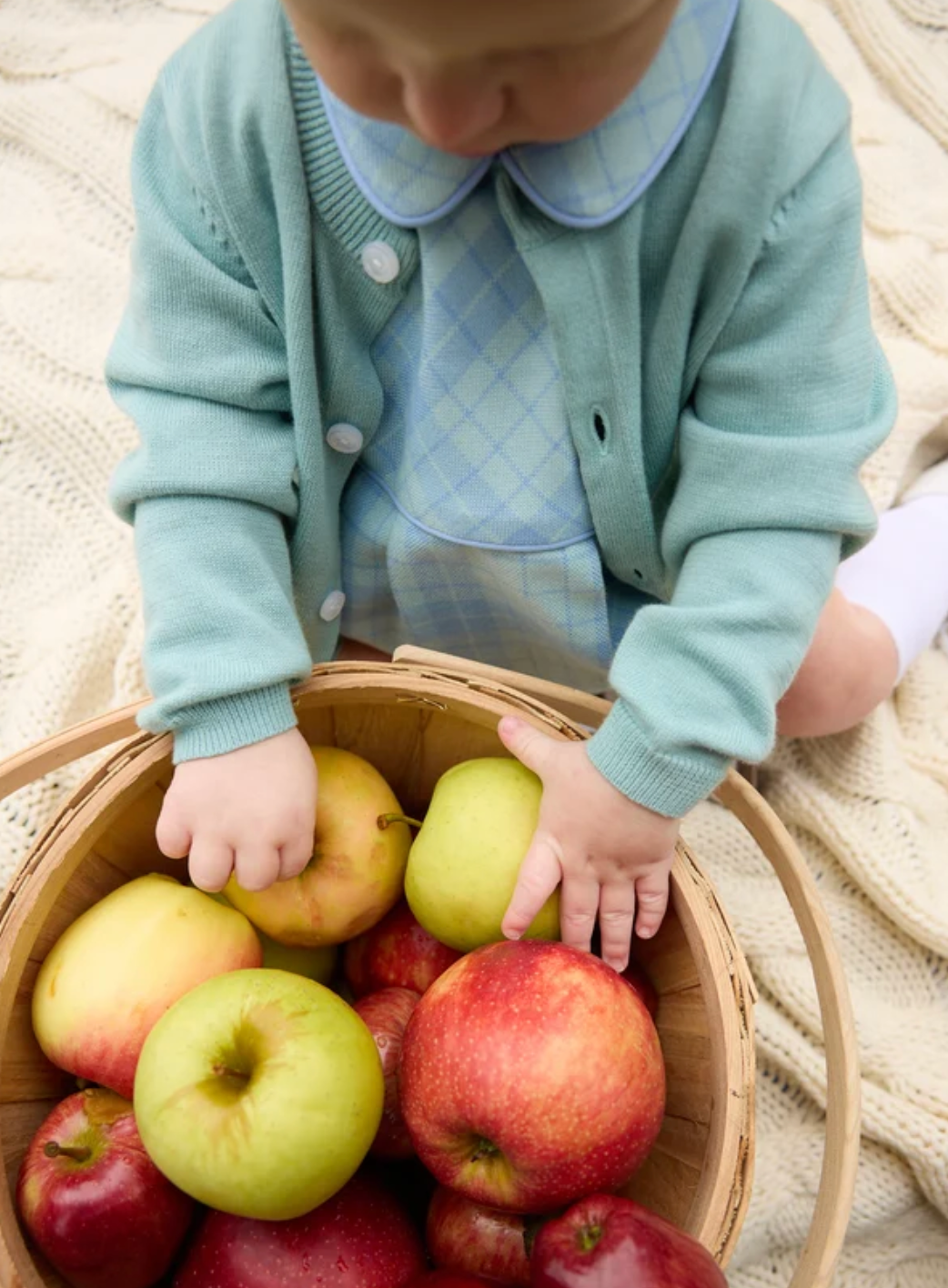 A baby wearing a light blue and white plaid outfit with a mint green cardigan reaches into a wooden basket filled with red and green apples, sitting on a cream-colored knit blanket.