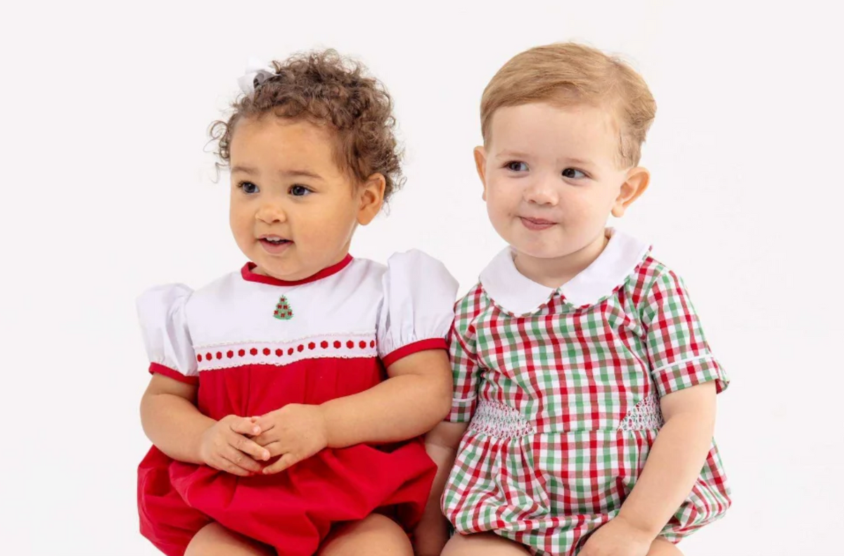 Two toddlers sitting side by side wearing matching holiday-themed dresses. The toddler on the left wears a red and white dress with white sleeves and a small green ornament detail.