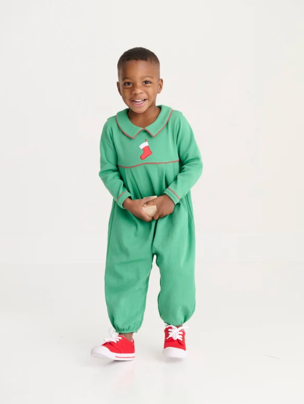 A young child wearing a festive green one-piece jumpsuit with a red and white stocking detail on the chest, paired with red sneakers, standing against a plain white studio background.