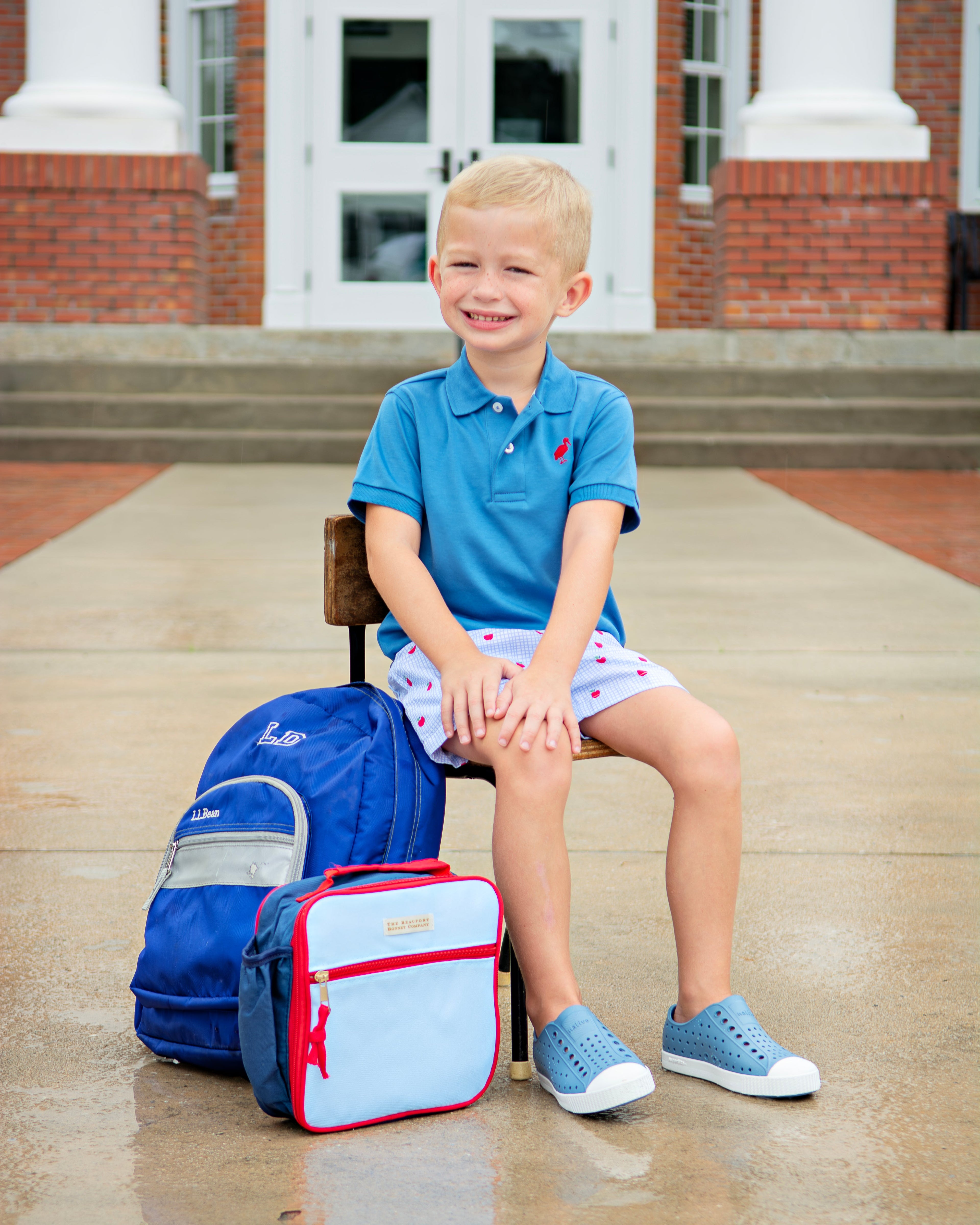 A young blond boy sits on a wooden chair in front of a brick building with white columns, wearing a blue polo shirt, patterned shorts, and blue shoes. A blue backpack and a light blue lunch bag with red trim sit beside him on the wet pavement.