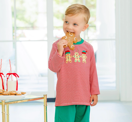 A young child wearing a red and white striped long-sleeve shirt with three gingerbread cookies on the chest and green pants, standing in a bright room and eating a gingerbread cookie.