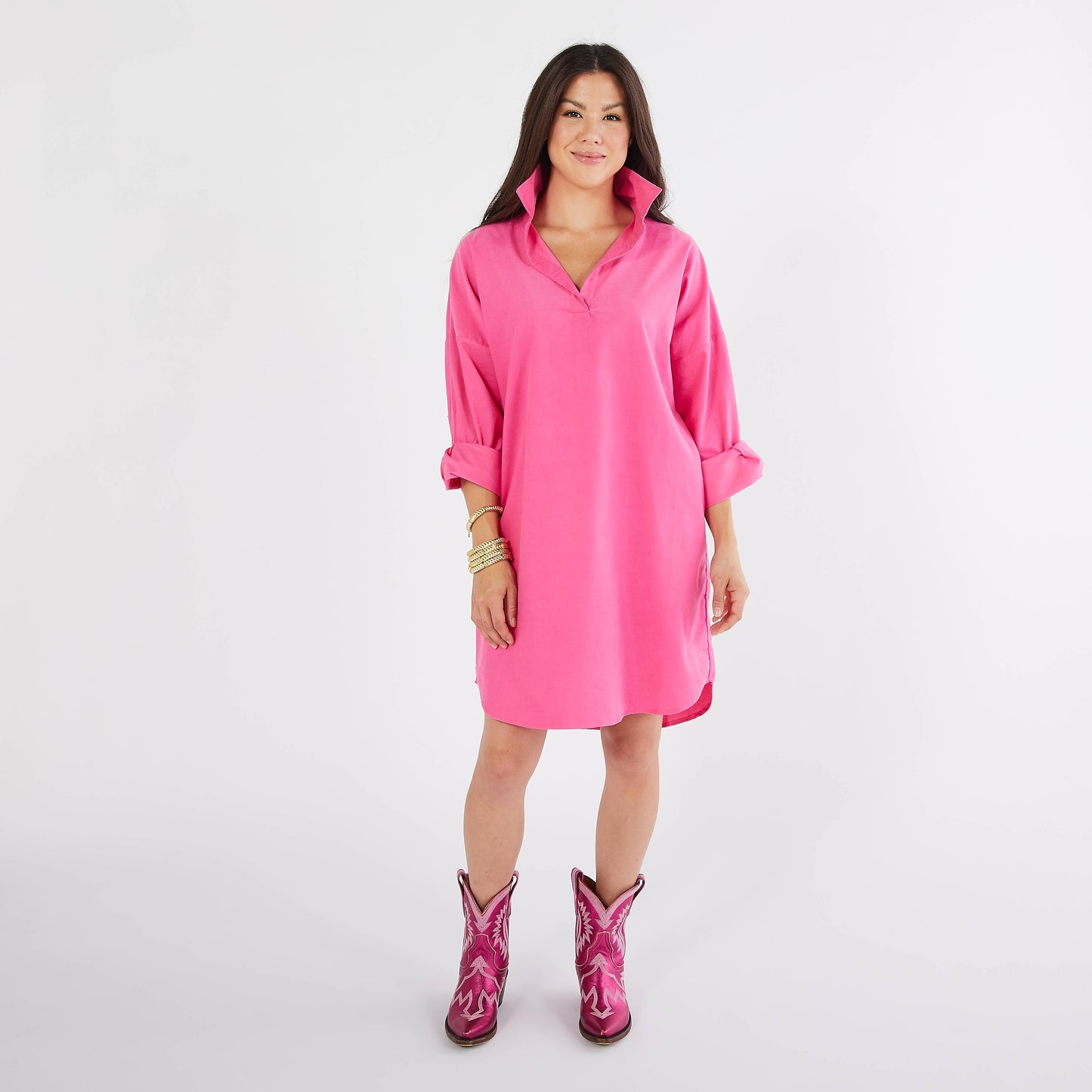 A woman modeling a bright pink oversized collared shirt dress with rolled sleeves, paired with matching pink cowboy boots, against a plain light gray studio background.