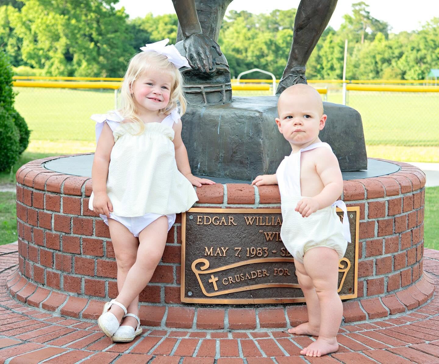 Two toddlers posing outdoors in front of a brick monument base with a bronze statue and a memorial plaque. The girl wears a white and yellow seersucker tie top diaper cover set and the boy wears a matching sunsuit.