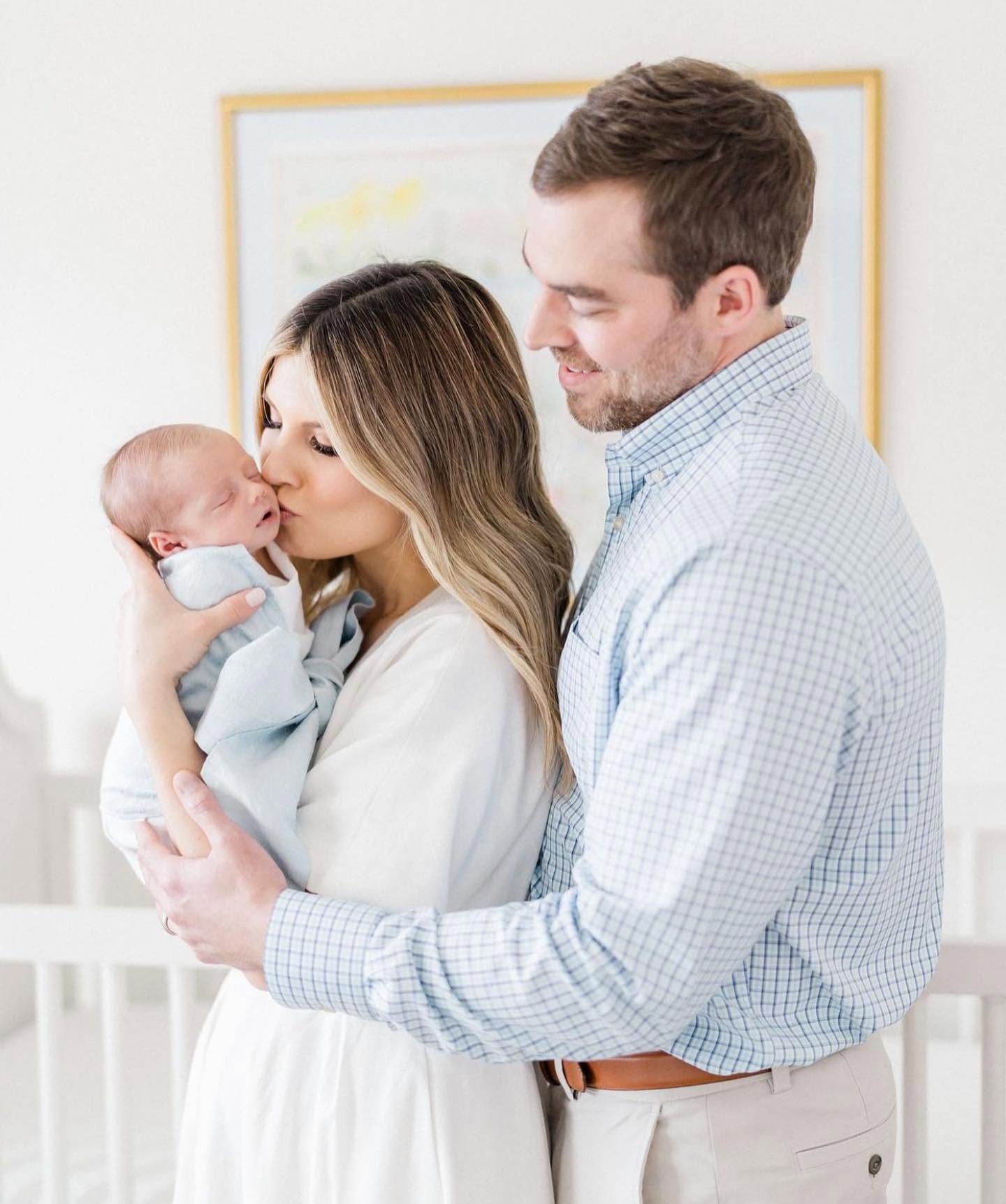 A family of three in a bright nursery setting, with a woman holding a sleeping newborn baby while kissing her husband on the cheek. The woman is wearing a white dress, and the man is wearing a light blue and white checkered button-down shirt and beige pants.