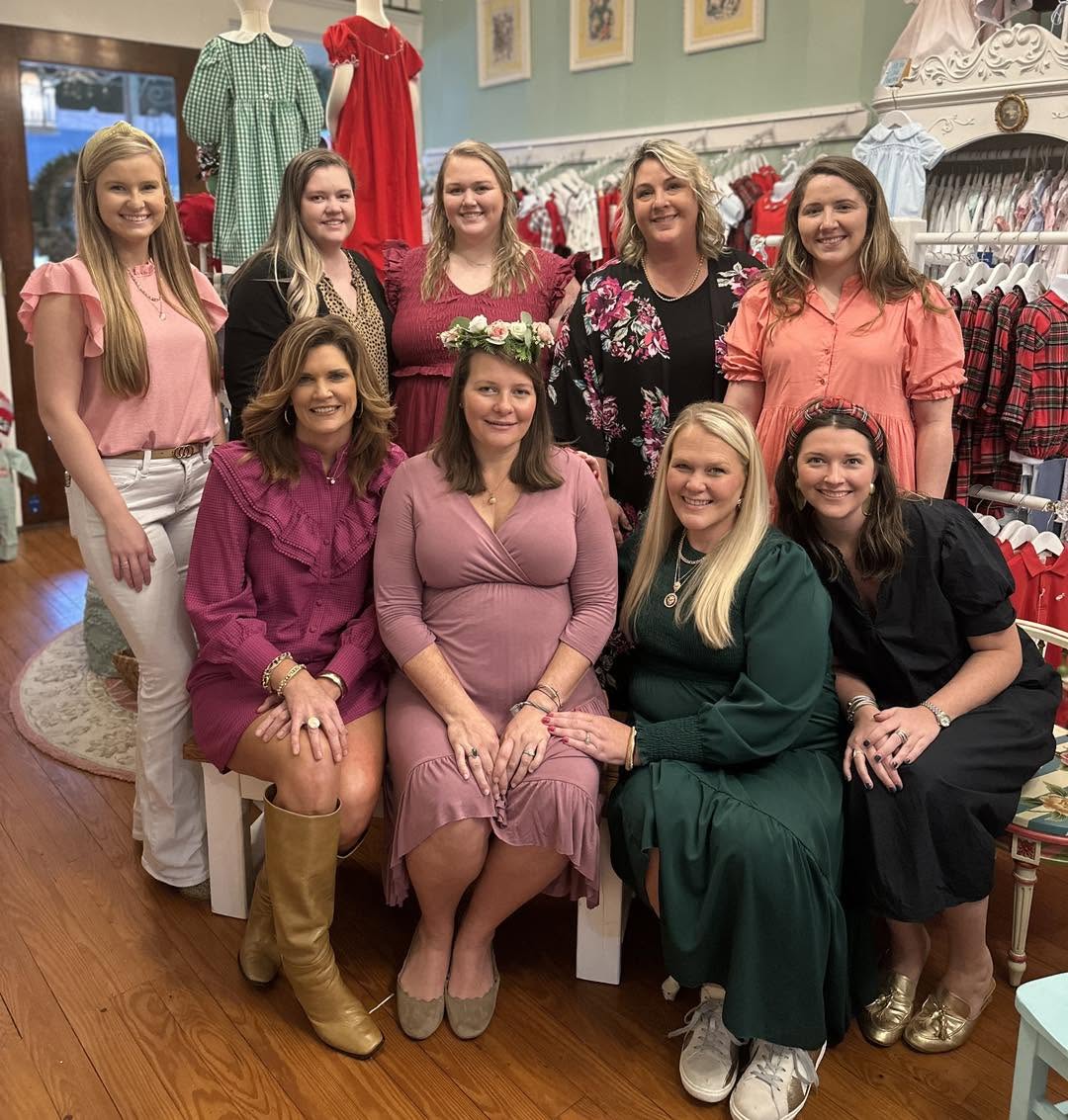 A group of nine women posing together inside a boutique clothing store celebrating an employee baby shower. Several women are wearing elegant dresses in shades of pink, green, and black, while others are dressed in casual outfits.
