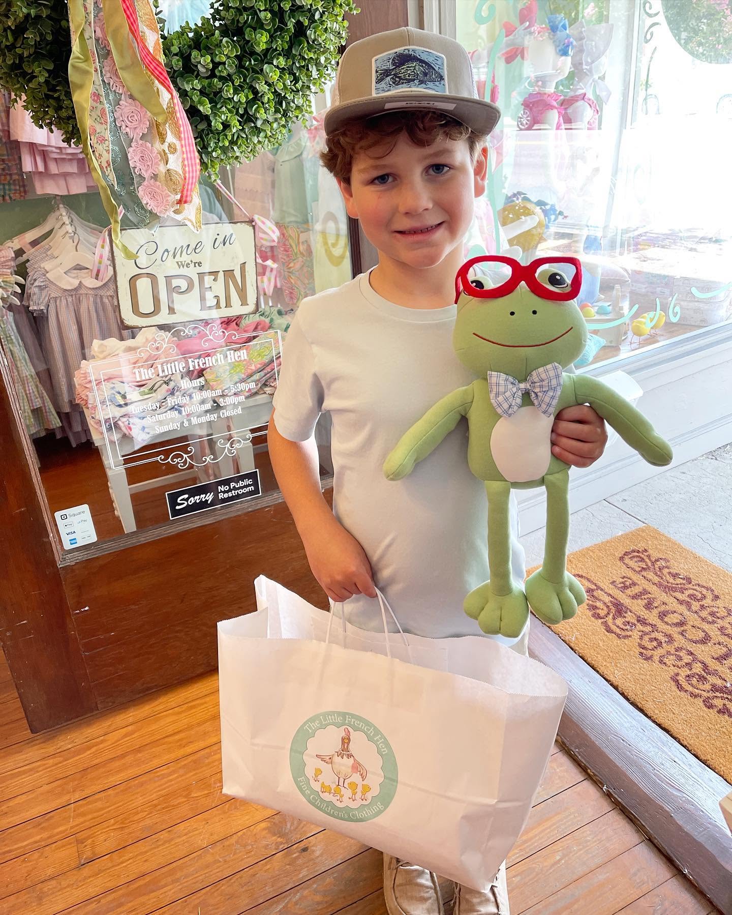 A young boy wearing a beige cap and white t-shirt stands in a shop holding a green plush frog toy with red glasses and a white shopping bag printed with the Little French Hen logo.