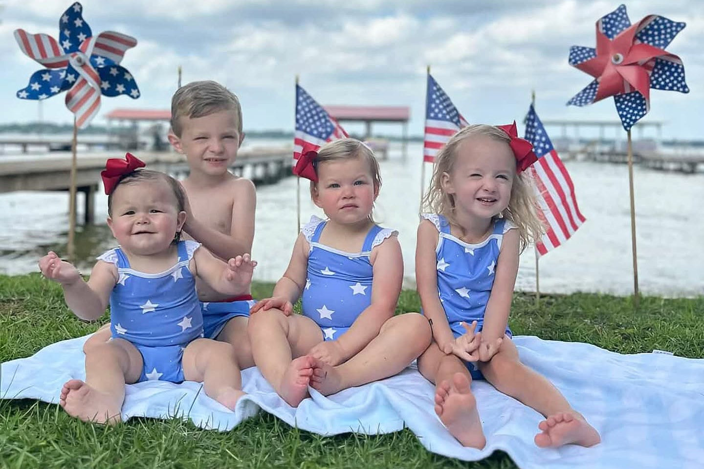 Four children wearing matching blue swimsuits with white stars sit on a white blanket on grass by the water at White Lake, NC, with American flags and pinwheel decorations in the background under a blue sky.