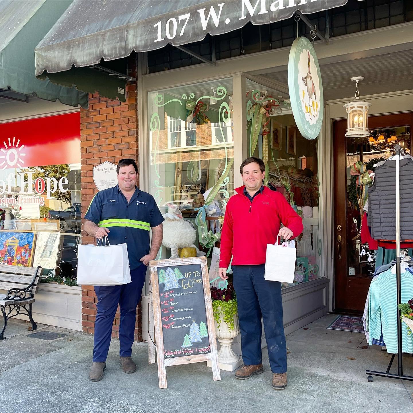 Two men standing on the sidewalk in front of the French Hen storefront, each holding white shopping bags. The man on the left wears a navy polo shirt with a yellow-green stripe and navy pants, while the man on the right wears a red long-sleeve shirt and dark pants.