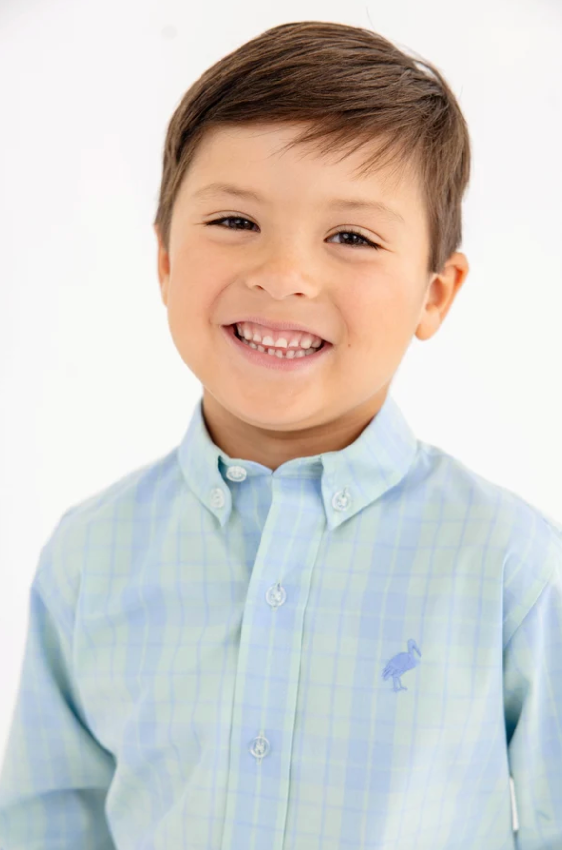 A young boy smiling, wearing a light blue and white checkered button-down shirt with a small blue stork logo on the chest, set against a plain white background.