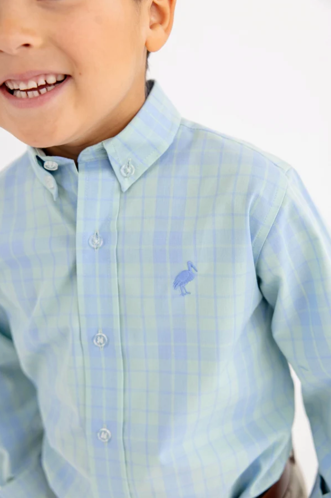 A close-up of a young boy wearing a light blue and mint plaid button-down shirt with a small embroidered stork logo on the chest.