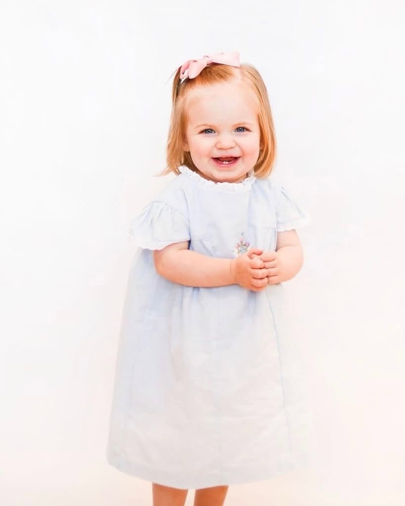 A smiling toddler girl with blonde hair and a pink bow in her hair, wearing a light blue short-sleeve dress with hand stitched cross and flower embroider, standing against a plain white background.