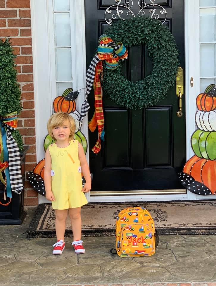 A young child wearing a yellow romper stands on a front porch next to a yellow patterned backpack. The porch features a black door decorated with a green wreath and autumn-themed decorations.