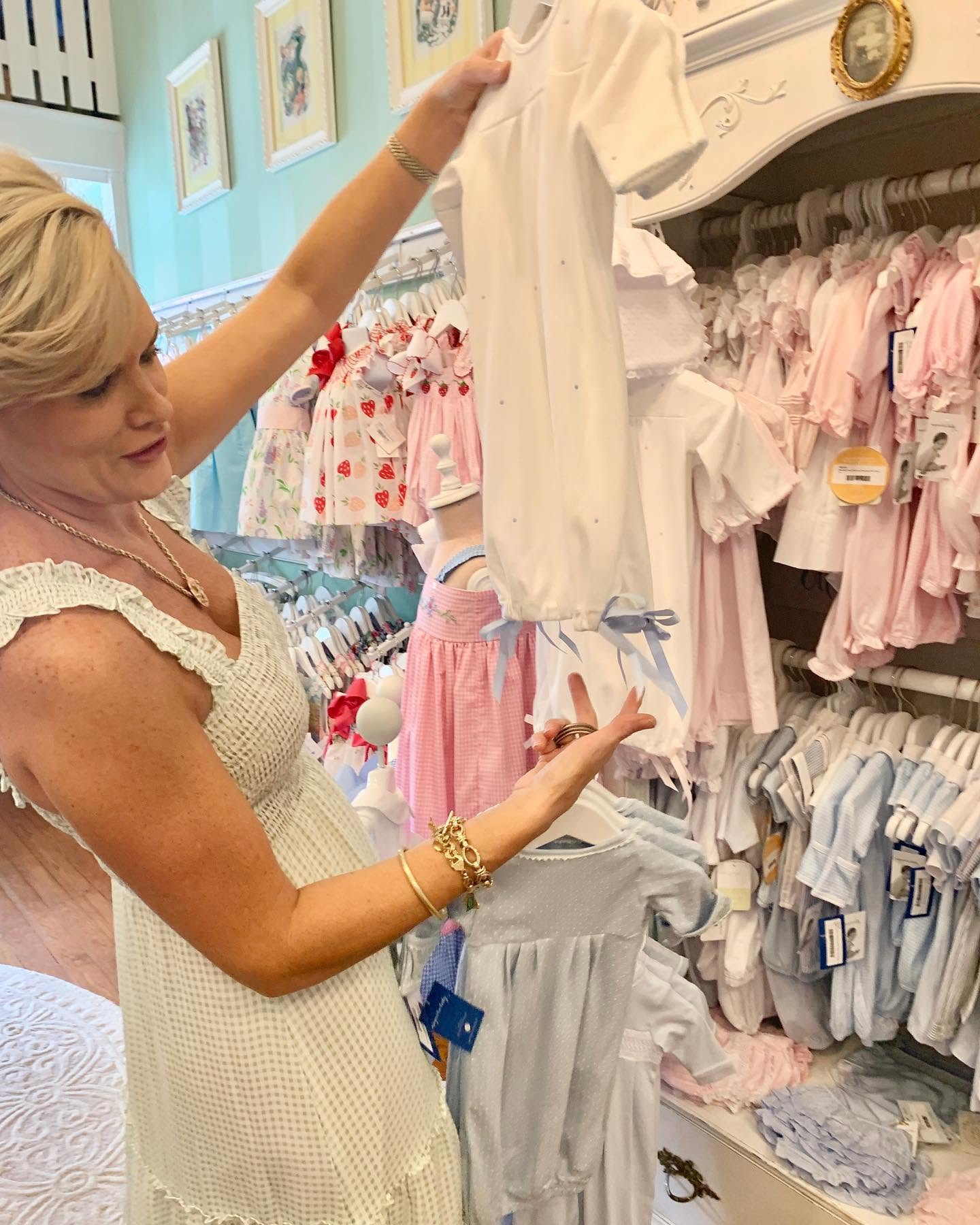 A woman in a white crochet dress browses baby clothing racks inside a boutique, holding up a white short-sleeve baby romper.