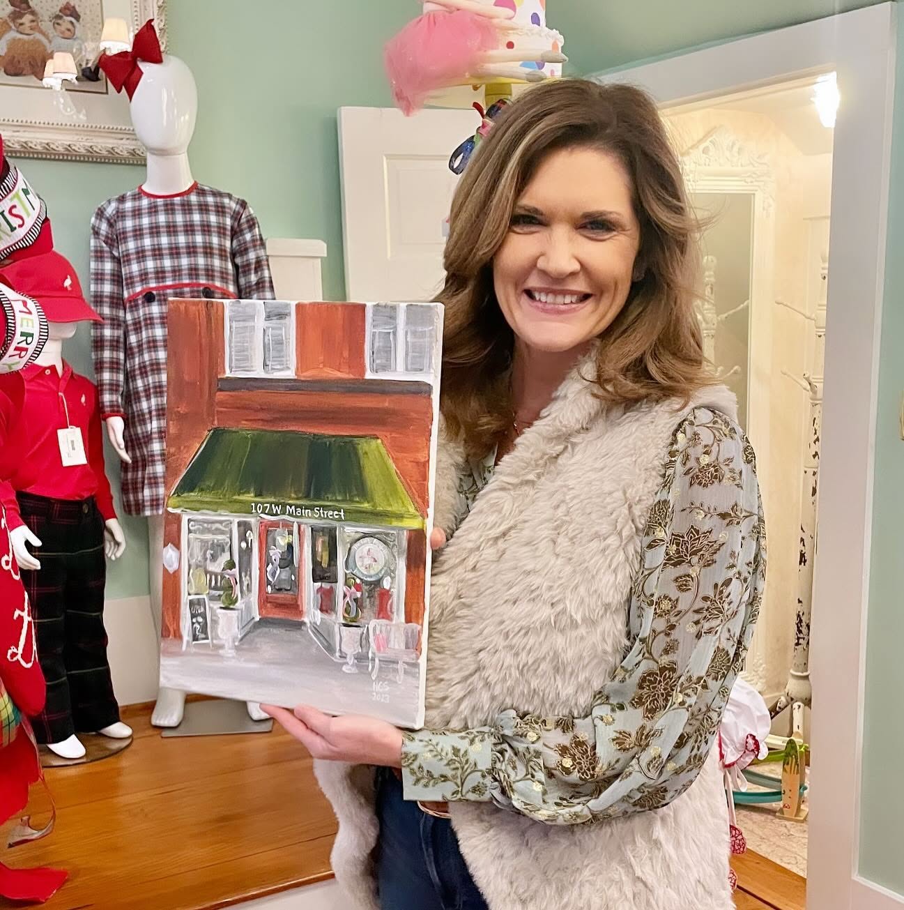 A smiling woman stands indoors holding a small painted storefront artwork. The room has light green walls, a doorway, mannequins in clothing, and colorful decor, making this a lifestyle shot rather than a clean product-only image.