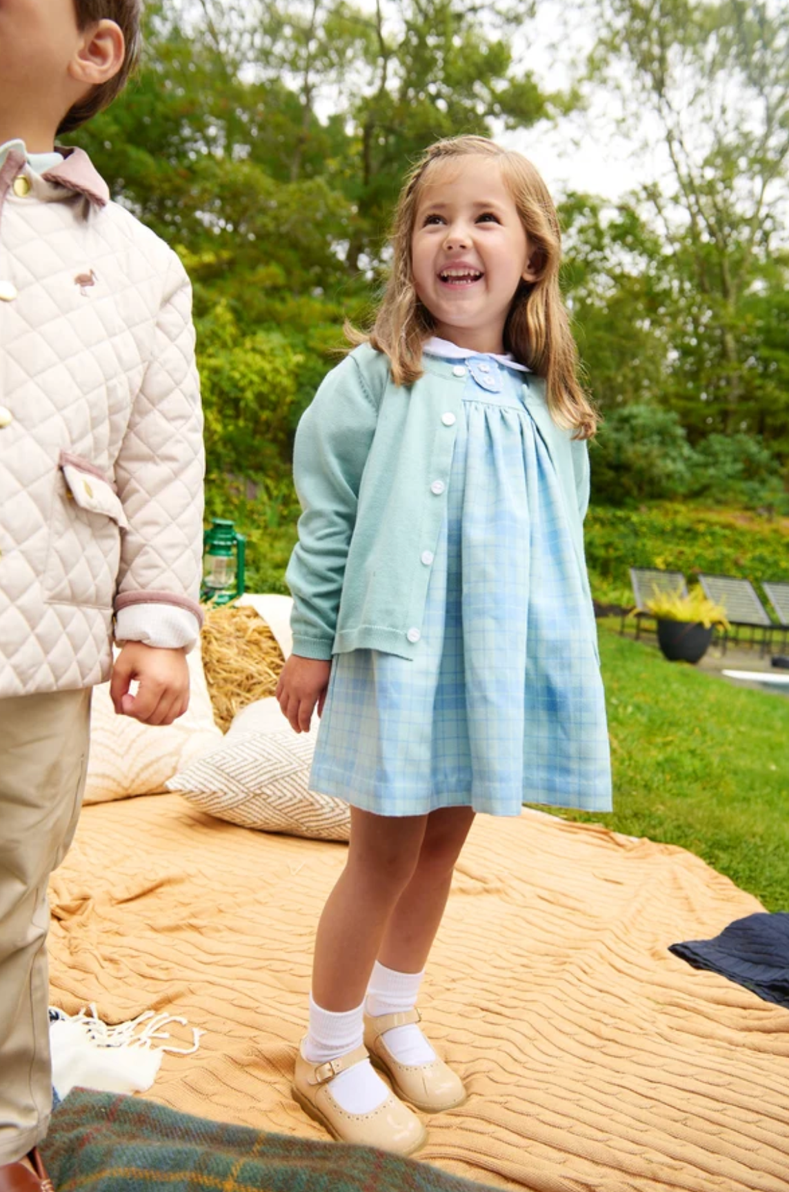 A young girl stands outdoors on a tan picnic blanket wearing a light blue plaid dress with a matching cardigan and white socks. Part of another child in a quilted jacket is visible at the left edge, with trees and patio furniture in the background.