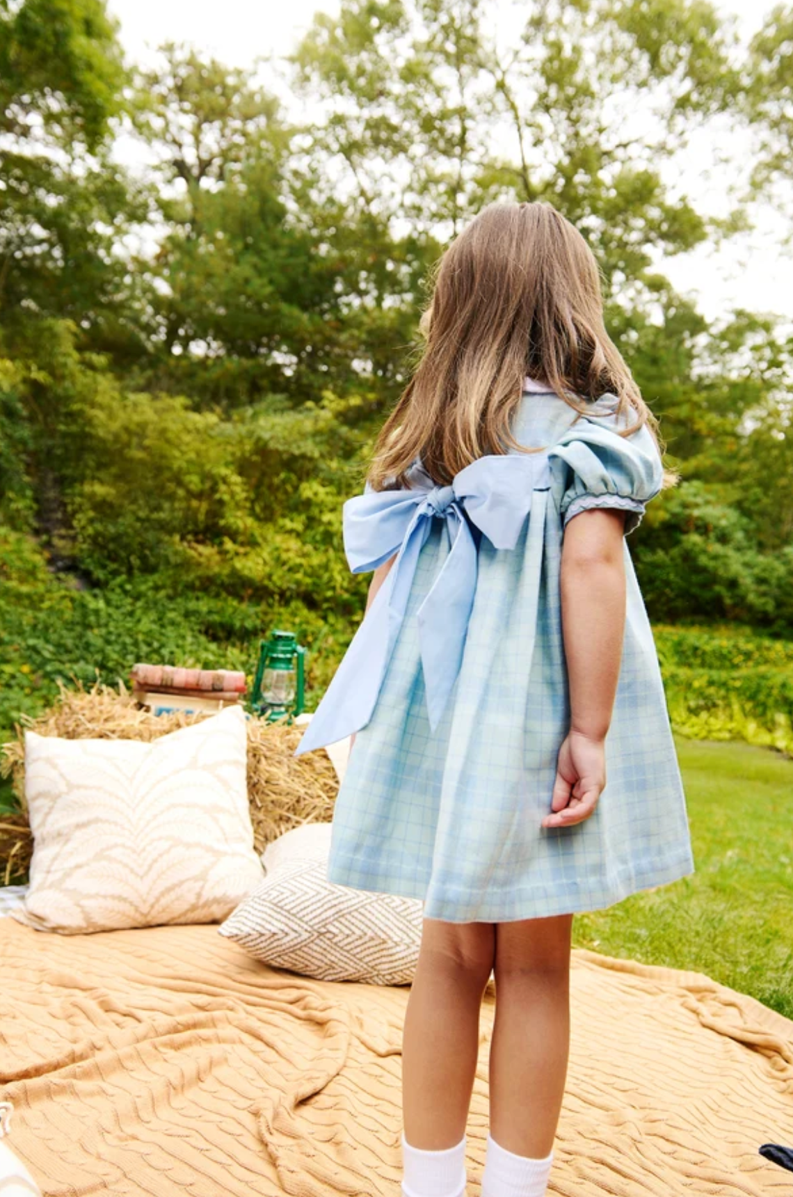 A young girl with long light brown hair stands outdoors on a beige blanket, viewed from behind. She is wearing a light blue plaid dress with a large bow on the back and puff sleeves.