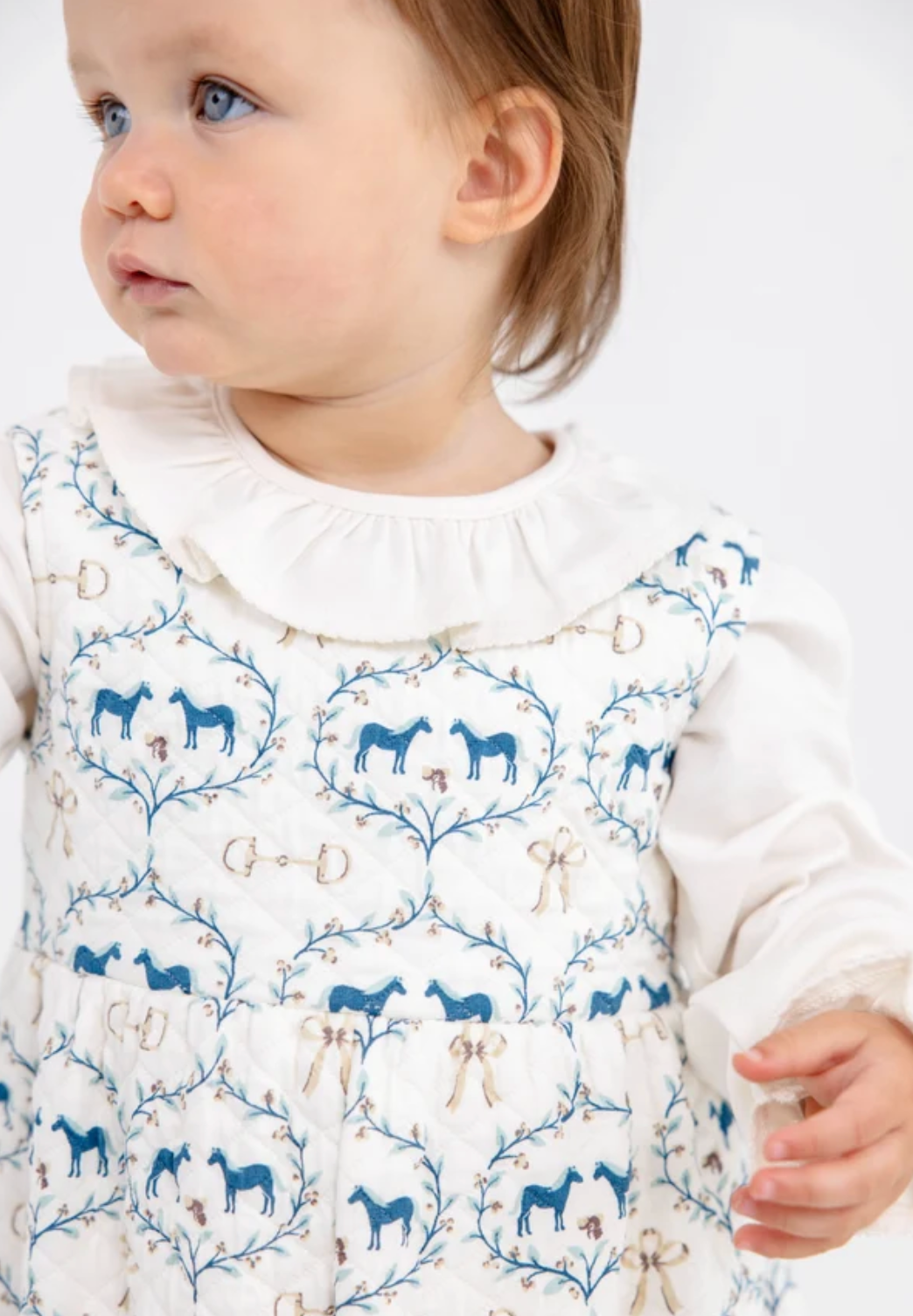 A close-up of a toddler wearing a white long-sleeve dress with a ruffled collar and an allover blue and beige horse-themed pattern, photographed against a plain light background.