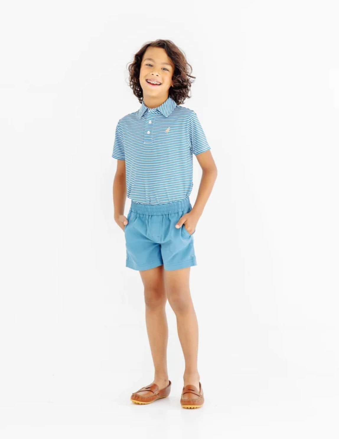 A young boy wearing a light blue and white striped short-sleeve polo shirt with matching light blue shorts, standing against a plain white studio background.