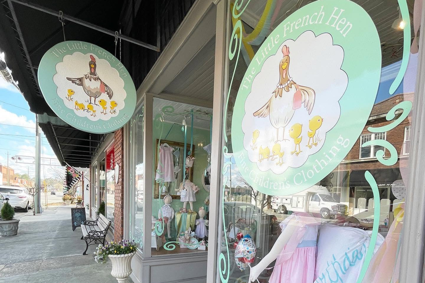 A storefront window display for 'The Little French Hen' nursery clothing, featuring pastel-colored baby clothes on mannequins and a large decorative sign on the window and a matching hanging sign above.