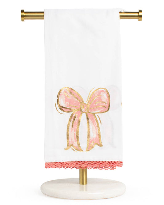 A white kitchen towel with a pink and gold bow design and a pink lace hem, hanging from a gold metal stand against a white background.